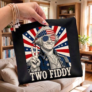 May include: A black fabric item featuring a graphic of George Washington wearing a stars and stripes bandana and sunglasses, making a peace sign. The background is a red, white, and blue sunburst pattern. The text TWO FIDDY is printed below.