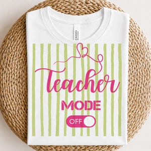 Puede incluir: Camiseta blanca con un diseño a rayas verdes y las palabras "Teacher MODE OFF" en rosa. La palabra "Teacher" está en cursiva y la palabra "MODE" en negrita. La camiseta está doblada.