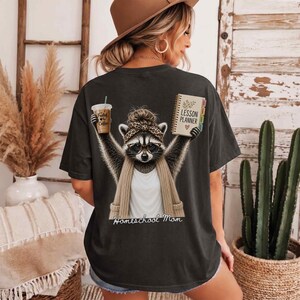 May include: A charcoal gray t-shirt featuring a graphic of a raccoon wearing a leopard print headband, glasses, and a beige cardigan. The raccoon holds a coffee cup labeled "Fueled by Coffee" and a "Lesson Planner" book. The text "Homeschool Mom" is below.