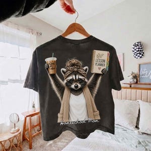May include: A charcoal gray t-shirt featuring a graphic of a raccoon wearing a leopard print headband, glasses, and a beige cardigan. The raccoon holds a coffee cup labeled Fueled by Coffee and a spiral-bound notebook titled Lesson Planner.