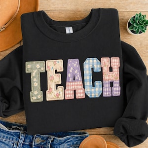 Może przedstawiać: Czarna bluza z napisem "TEACH" w kolorowych literach w stylu patchwork. Litery mają kwiatowe, kratowe i wzorzyste wzory w odcieniach zieleni, różu, błękitu i fioletu. Bluza jest złożona na drewnianej powierzchni.