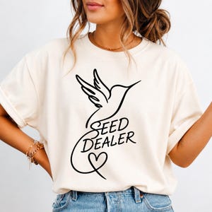 Könnte beinhalten: Cremefarbenes T-Shirt mit einer schwarzen Strichzeichnung eines Kolibris, dem Text "SEED DEALER" und einem Herz. Das Design ist mittig auf der Vorderseite. Das T-Shirt hat hochgekrempelte Ärmel.