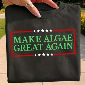 Könnte beinhalten: Ein schwarzes Sweatshirt mit Rundhalsausschnitt, das einen roten rechteckigen Rahmen mit fünf weißen Sternen über und unter dem Text "MAKE ALGAE GREAT AGAIN" in grünen Großbuchstaben auf der Vorderseite des Kleidungsstücks zeigt.