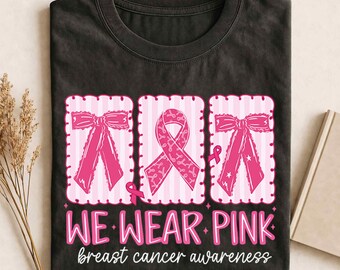 May include: A black t-shirt featuring a graphic with three vertical rectangular panels. The outer panels show pink bows, and the center panel displays a pink awareness ribbon. Below, the text reads WE WEAR PINK breast cancer awareness.