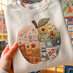 May include: A cream-colored crewneck sweatshirt featuring a large, central apple-shaped embroidery. The apple is divided into seasonal quilt-like sections displaying flowers, a birdhouse, a sunflower, strawberries, autumn leaves, a pumpkin, a basket, snowflakes, a winter hat, mittens, a cabin, and a pine tree.
