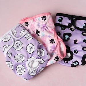 Könnte beinhalten: Drei wiederverwendbare Windeln für Haustiere in verschiedenen Mustern. Eine ist lavendelfarben mit weißen Hundeillustrationen, eine andere ist rosa mit Pfotenabdrücken und die dritte ist lavendelfarben mit schwarzen und teal Leopardenflecken. Alle haben weiße Verzierungen.