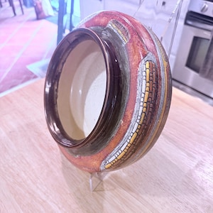 May include: A round ceramic bowl featuring a dark brown interior and a textured exterior rim. The outer surface displays a mottled reddish-orange glaze with decorative vertical bands of yellow and silver brick-like patterns encircling the bowl.