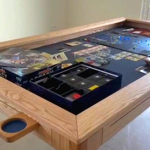 Op de afbeelding: Een grote houten speeltafel met een donker speeloppervlak. De tafel heeft een lichtbruine, eikenhouten afwerking en bevat een ingebouwde bekerhouder. Speldozen en stukken zijn zichtbaar, wat wijst op een bordspelopstelling.