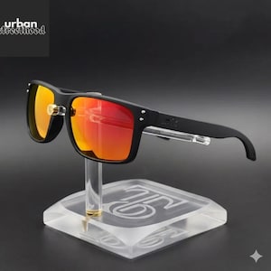 Puede incluir: Gafas de sol negras con lentes naranja-rojas se exhiben en un soporte transparente. Las gafas tienen una montura rectangular y un peque&ntilde;o logotipo en la patilla. El soporte tiene un acento dorado y un logotipo en la base. El texto "urban streetfood" est&aacute; en la esquina superior izquierda.