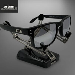 May include: Black sunglasses with grey lenses on a black and silver stand. The sunglasses have a square frame and the Oakley logo. The stand has a mechanical design, and the text "urban street hood" is in the upper left corner.