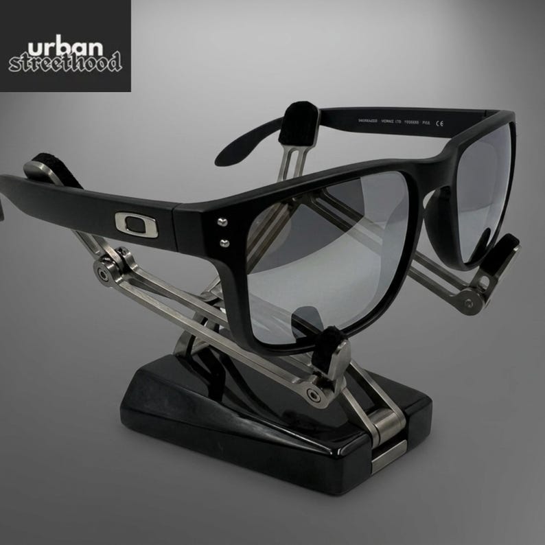 May include: Black sunglasses with grey lenses on a black and silver stand. The sunglasses have a square frame and the Oakley logo. The stand has a mechanical design, and the text "urban street hood" is in the upper left corner.