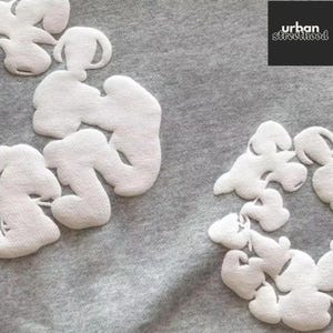 May include: Close-up of a gray fabric with white, cloud-shaped appliqu&eacute;s. The appliqu&eacute;s are clustered in groups across the fabric. The upper right corner features a black square with the text "urban streetthood" in white.