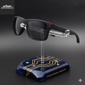 May include: Black sunglasses with dark lenses and red detailing are displayed on a clear stand. The stand is mounted on a blue base with a gold circuit board design. The text "TG Smart Collection - Obsidian Ed." is visible.