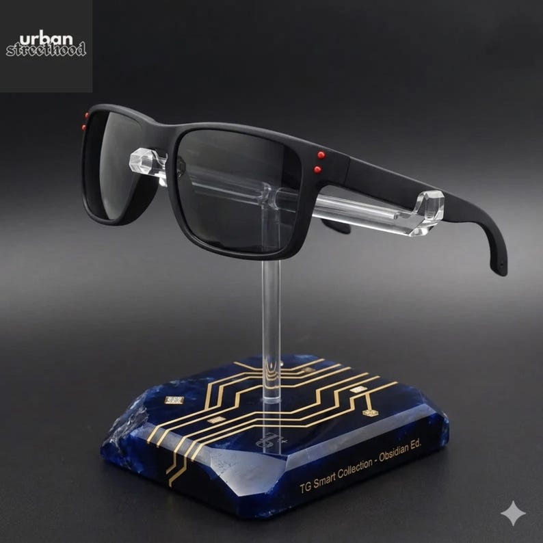 May include: Black sunglasses with dark lenses and red detailing are displayed on a clear stand. The stand is mounted on a blue base with a gold circuit board design. The text "TG Smart Collection - Obsidian Ed." is visible.