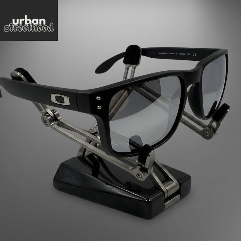 Puede incluir: Gafas de sol negras con lentes grises expuestas en un soporte negro y plateado. Las gafas tienen una montura cuadrada y el logotipo de Oakley es visible. El soporte tiene un dise&ntilde;o mec&aacute;nico. El texto "urban street hood" es visible.