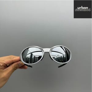 Puede incluir: Gafas de sol con montura plateada y lentes ovaladas. Las lentes reflejan el entorno. Las gafas de sol tienen un diseño elegante y moderno. La marca "OKLEY" es visible en la montura. La imagen también incluye el texto "urban street hood".
