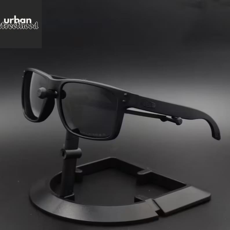 Puede incluir: Gafas de sol negras con montura cuadrada expuestas sobre un soporte negro. Las gafas de sol tienen lentes oscuras y un acabado mate. El texto "urban streetthood" est&aacute; en la esquina superior izquierda.