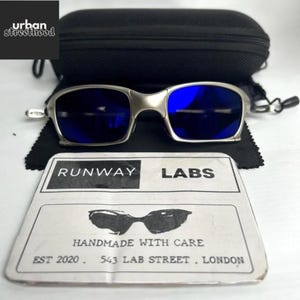 May include: Silver-framed sunglasses with blue lenses, resting on a black cloth. A black case and a card with the text "RUNWAY LABS" and "HANDMADE WITH CARE" are also visible. The sunglasses have a modern, sleek design.