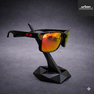 May include: Black sunglasses with orange and yellow mirrored lenses are displayed on a black geometric stand. The sunglasses have a black frame with red accents. The stand has a faceted base and a slender support.