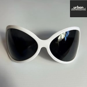 Puede incluir: Gafas de sol blancas con una montura audaz en forma de mariposa y lentes oscuras. Diseño moderno. El texto "urban streetthood" está en la esquina superior derecha.