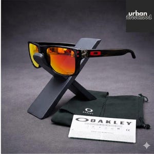 May include: Black Oakley sunglasses with orange mirrored lenses are displayed on a gray stand. The sunglasses have a rectangular frame with red accents. A black drawstring bag and a white card with the Oakley logo are also present.