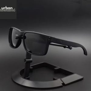 May include: Black sunglasses with a square frame are displayed on a black stand. The sunglasses have dark lenses and a matte finish. The text "urban streetthood" is in the upper left corner.