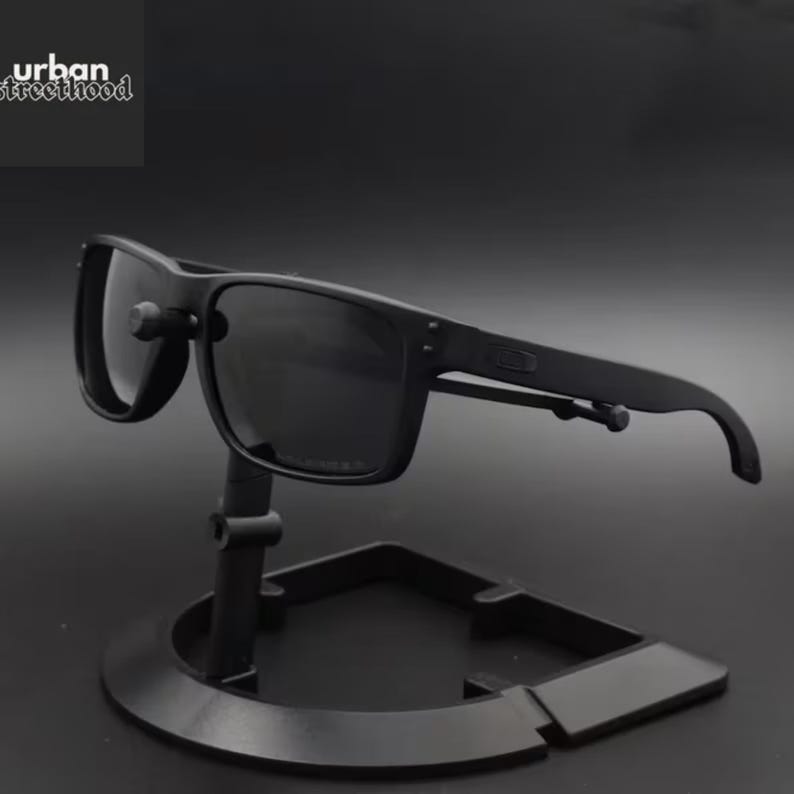 May include: Black sunglasses with a square frame are displayed on a black stand. The sunglasses have dark lenses and a matte finish. The text "urban streetthood" is in the upper left corner.