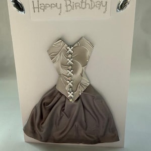 May include: A handmade birthday card featuring a 3D dress design. The dress is made from silver and grey fabric, with white ribbon detailing. The card has the words "Happy Birthday" in silver glitter and is decorated with rhinestones.
