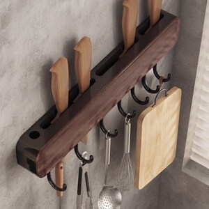 Op de afbeelding: Een wandgemonteerde keukenorganizer met een donkerhouten horizontale balk met sleuven voor vier messen. De unit bevat vijf zwarte metalen haken waaraan een soeplepel, metalen tang, schuimspaan, garde en een houten snijplank hangen.