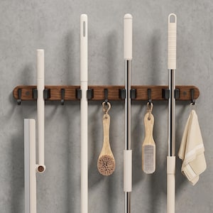 May include: A wall-mounted wooden storage rack featuring multiple black clips and hooks. The rack holds various cleaning tools, including long-handled mops, brushes with wooden handles, and a folded beige cloth hanging from a hook on the right side.
