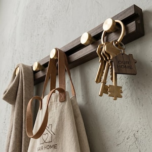 May include: A dark wood wall-mounted key and coat rack with brass hooks. A beige tote bag with brown leather straps hangs from a hook, along with a beige scarf. A set of keys with a wooden "OUR HOME" tag is also hanging.