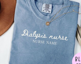 Puede incluir: Una camiseta azul vaquero con las palabras "Dialysis nurse" y "Nurse Name" en escritura blanca. Un collar dorado con un colgante descansa sobre la camisa. Un sombrero marrón y una planta seca están en el fondo.