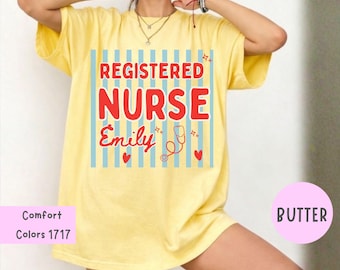Puede incluir: Una camiseta de color amarillo mantequilla con un gráfico de rayas verticales azul claro. El texto REGISTERED NURSE aparece en letras rojas, con el nombre Emily en cursiva roja debajo. Un estetoscopio rojo y corazones rojos completan el diseño.