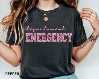 Puede incluir: Camiseta gris oscuro con las palabras "department EMERGENCY" en rosa en escritura cursiva y letras de bloque. Las letras de bloque tienen un patrón de camuflaje rosa. La palabra "pepper" está en la parte inferior de la camiseta.