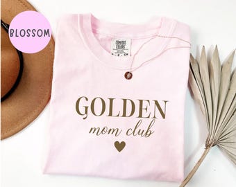 Puede incluir: Una camiseta de manga corta de color rosa claro con el texto GOLDEN mom club en letras de color marrón oscuro y un pequeño icono de corazón de color marrón oscuro debajo del texto. La camiseta tiene cuello redondo y etiqueta Comfort Colors.
