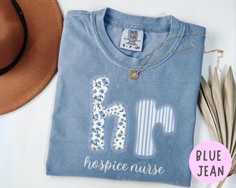 Puede incluir: Una camiseta azul vaquera Comfort Colors con las palabras "hospice nurse" en escritura blanca. Las letras "h" y "r" están aplicadas con patrones florales y a rayas. Un collar dorado con un colgante descansa sobre la camiseta.