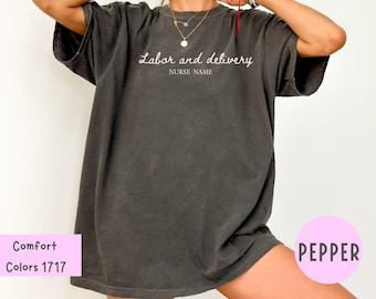 Puede incluir: Una camiseta gris oscuro con las palabras "Labor and delivery" y "Nurse Name" en escritura blanca. La camiseta es de color confort y tiene un ajuste relajado. El color está etiquetado como "Pepper".