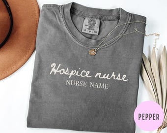 Puede incluir: Una camiseta gris Comfort Colors con las palabras "Hospice nurse" y "Nurse Name" en escritura blanca. Un collar dorado con un colgante descansa sobre la camisa. Un sombrero marrón y plantas secas están en el fondo.