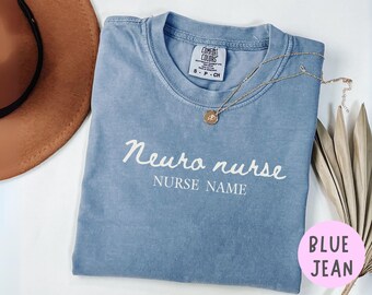 Puede incluir: Una camiseta azul vaquero Comfort Colors con las palabras "Neuro nurse" y "Nurse Name" en escritura blanca. Un collar dorado con un colgante descansa sobre la camisa. Un sombrero marrón y una planta seca están en el fondo.