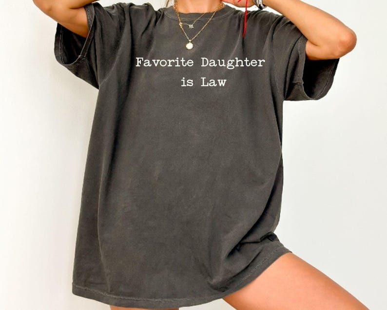 Puede incluir: Camiseta gris oscuro de gran tama&ntilde;o con el texto blanco "Favorite Daughter is Law". La camiseta tiene mangas cortas y un corte relajado, adecuada para uso casual. La tela parece suave y ligeramente descolorida.