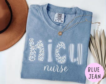 Puede incluir: Una camiseta azul vaquero Comfort Colors con las palabras "NICU nurse" en blanco. Las letras "N", "I" y "U" están decoradas con un estampado floral azul. Un collar de oro con un colgante descansa sobre la camiseta.