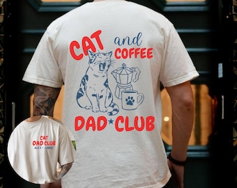 Puede incluir: Camiseta color crema con el texto "CAT and COFFEE DAD CLUB" en rojo y azul. El diseño incluye un gato azul, una cafetera y una huella de pata en una taza. La camiseta es de manga corta.