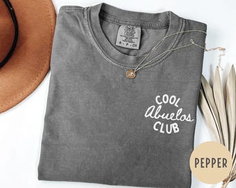 Puede incluir: Una camiseta gris Comfort Colors con las palabras "COOL Abuelos CLUB" en escritura blanca. Un collar dorado con un colgante descansa sobre la camiseta. La camiseta está doblada y tiene una etiqueta de color "Pepper".