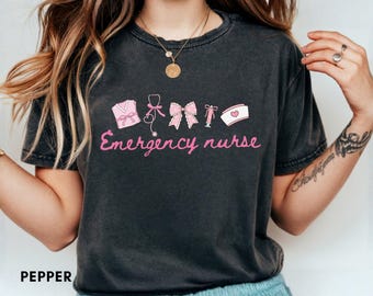 Puede incluir: Camiseta gris oscuro con la frase "Emergency nurse" en rosa. La camiseta presenta ilustraciones rosas de símbolos médicos, incluyendo un uniforme de enfermera, estetoscopio, lazo, jeringa y gorro de enfermera. La palabra "PEPPER" está impresa en la parte inferior.