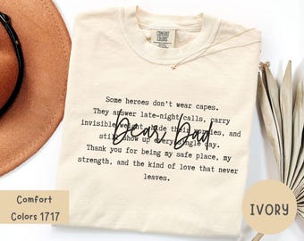 Puede incluir: Camiseta de color marfil con la frase "Dear Dad" en fuente manuscrita. La camiseta presenta un mensaje impreso sobre héroes y agradecimiento. La camiseta Comfort Colors 1717 se muestra con un sombrero marrón y elementos decorativos.