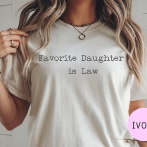 Puede incluir: Camiseta de color marfil con el texto "Favorite Daughter is Law" en una fuente sencilla sin serifa. La camiseta tiene mangas cortas y cuello redondo. Un c&iacute;rculo rosa con la palabra "IVORY" est&aacute; en la esquina inferior derecha.