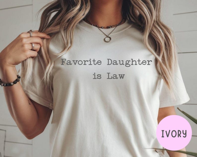 Puede incluir: Camiseta de color marfil con el texto "Favorite Daughter is Law" en una fuente sencilla sin serifa. La camiseta tiene mangas cortas y cuello redondo. Un c&iacute;rculo rosa con la palabra "IVORY" est&aacute; en la esquina inferior derecha.