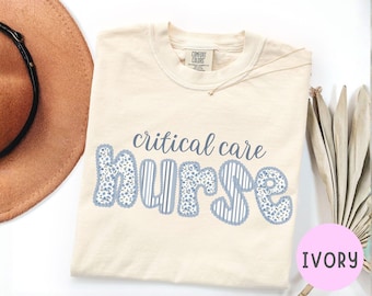 Puede incluir: Camiseta de color marfil con las palabras "critical care nurse" en azul. La palabra "nurse" está en una fuente decorativa con patrones florales y a rayas. La camiseta es de la marca Comfort Colors.