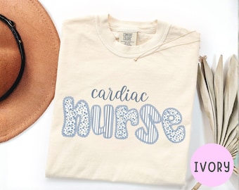 Puede incluir: Camiseta de color marfil con las palabras "cardiac nurse" en letras azules. La palabra "cardiac" está en una fuente cursiva, y "nurse" está en una fuente lúdica con un diseño floral y a rayas. La camiseta es de la marca Comfort Colors.