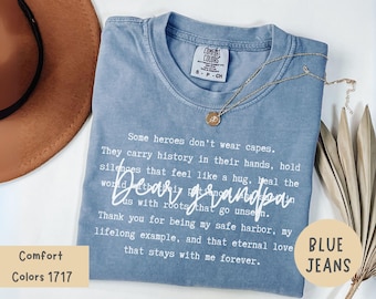 Puede incluir: Una camiseta azul Comfort Colors con el texto "Dear Grandpa" y un collar dorado. La camiseta también tiene el texto "Some heroes don't wear capes" y "Blue Jeans".